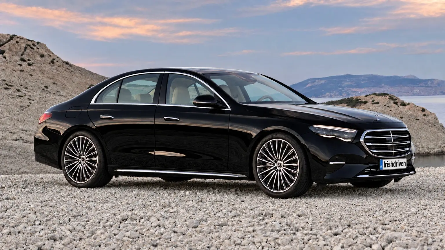 A black Mercedes-Benz E-Class saloon parked on a pebble shore by the sea