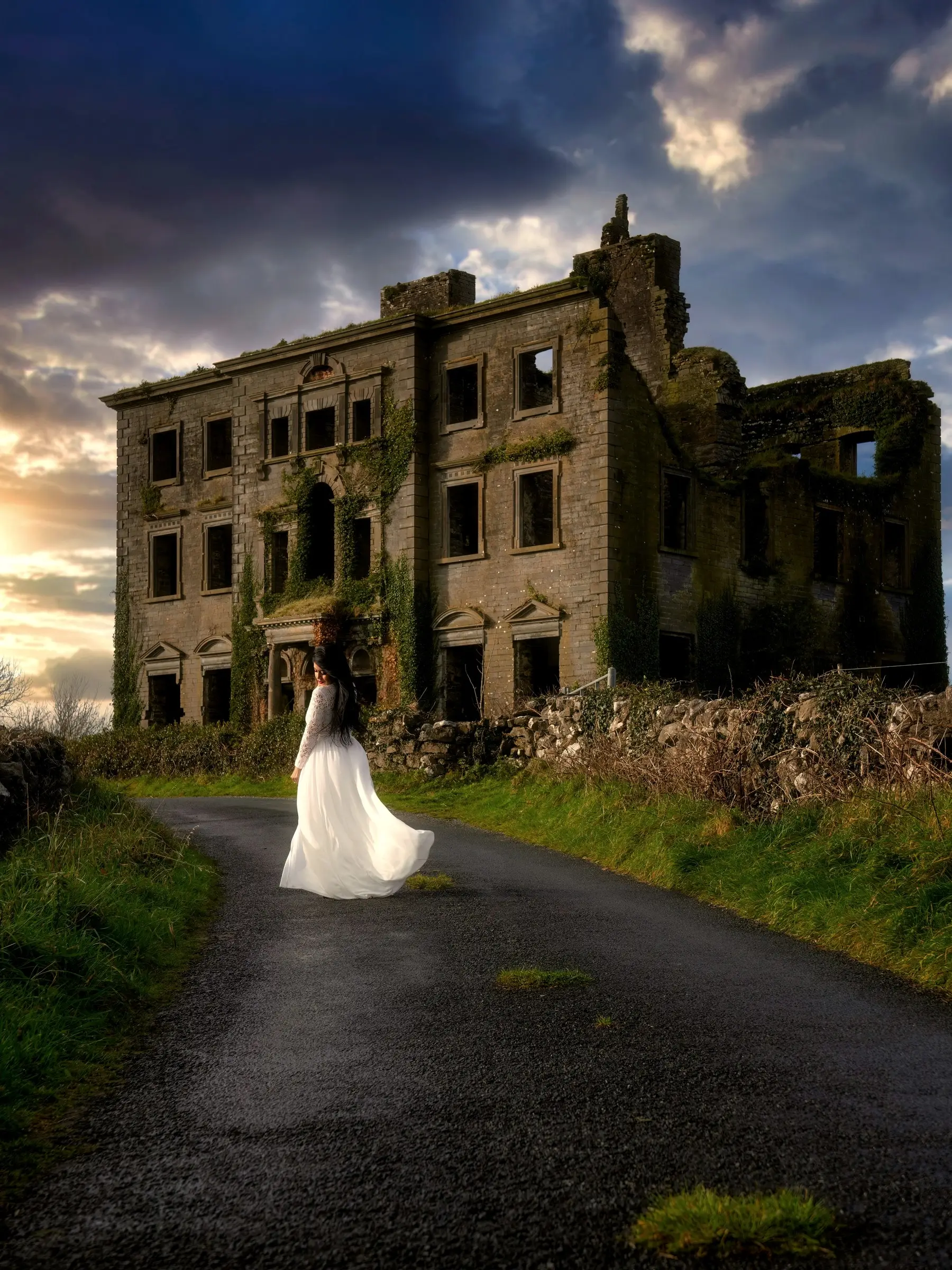 A bride in a flowing white dress on a country lane below an old ivy-covered Irish house at sunset